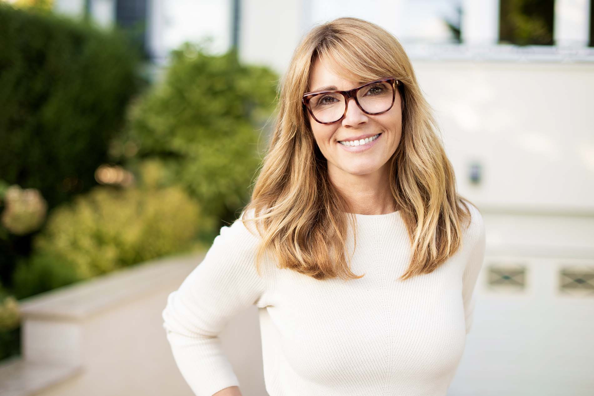 Woman in front of house is white sweater enjoying her breast augmentation without an implant.